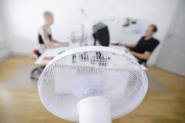 Im Vordergrund ist ein weißer Ventilator von hinten zu sehen und im Hintergrund unscharf zu erkennen sind zwei Personen, die sich an einem Schreibtischen gegenübersitzen